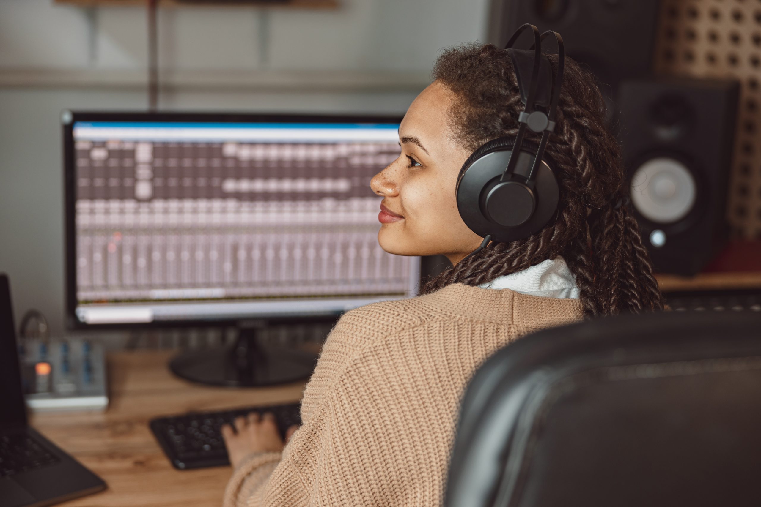 African American female musician, producer, sound engineer in headphones sits at a digital audio workstation software control panel while recording a new track for an album in a soundproof room