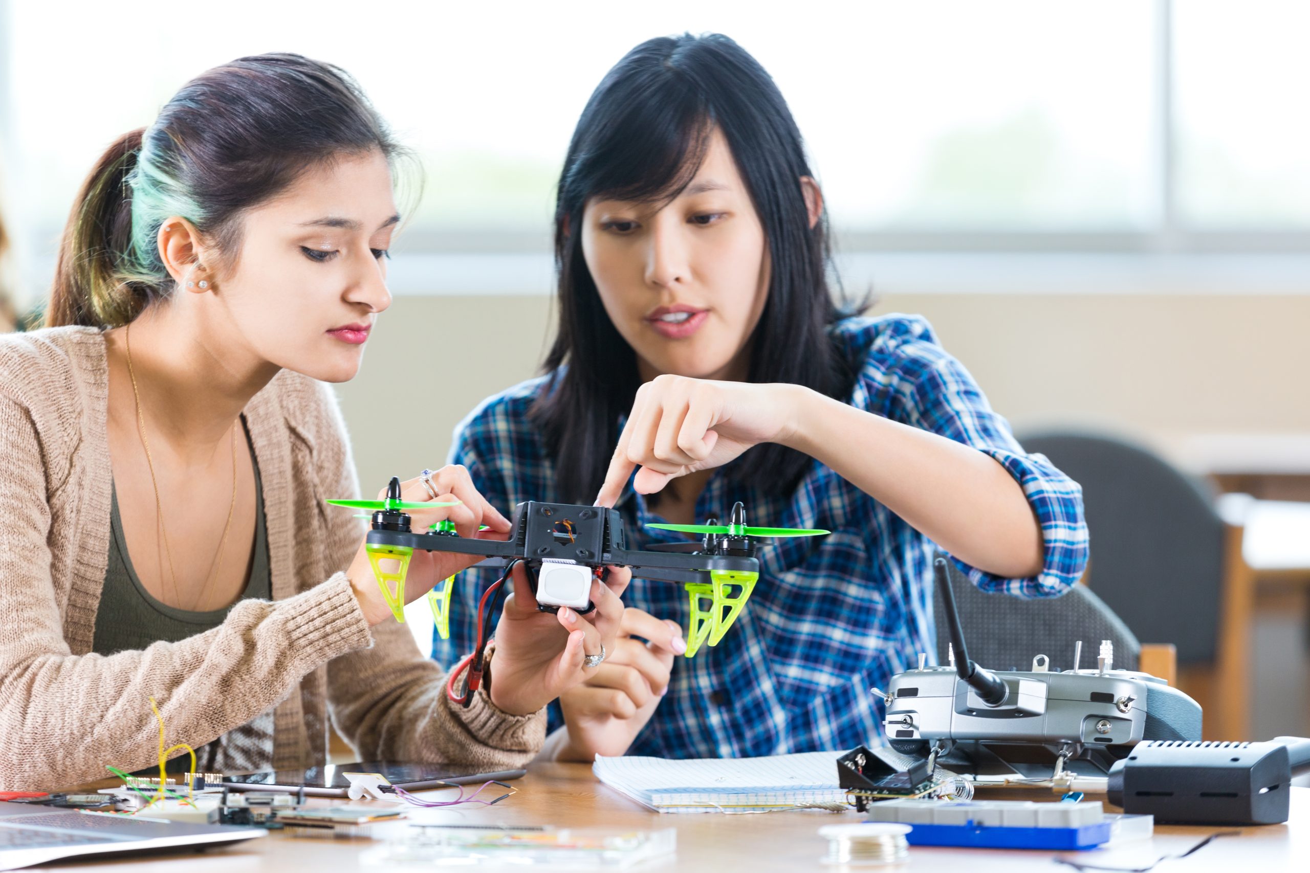 Pretty Asian teacher at STEM school helps Hispanic student with drone in engineering class. The teacher is pointing to something on the drone as the student looks on. The controller and other parts or tools are on the table. The teacher is wearing a blue plaid shirt and the student is wearing trendy layered clothing.